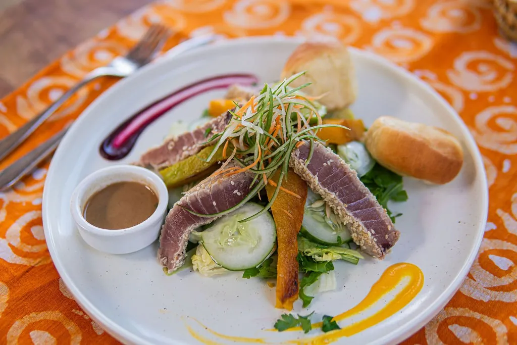 Slices of seared tuna with salad, a dipping sauce and bread on a white plate over a patterned orange cloth.