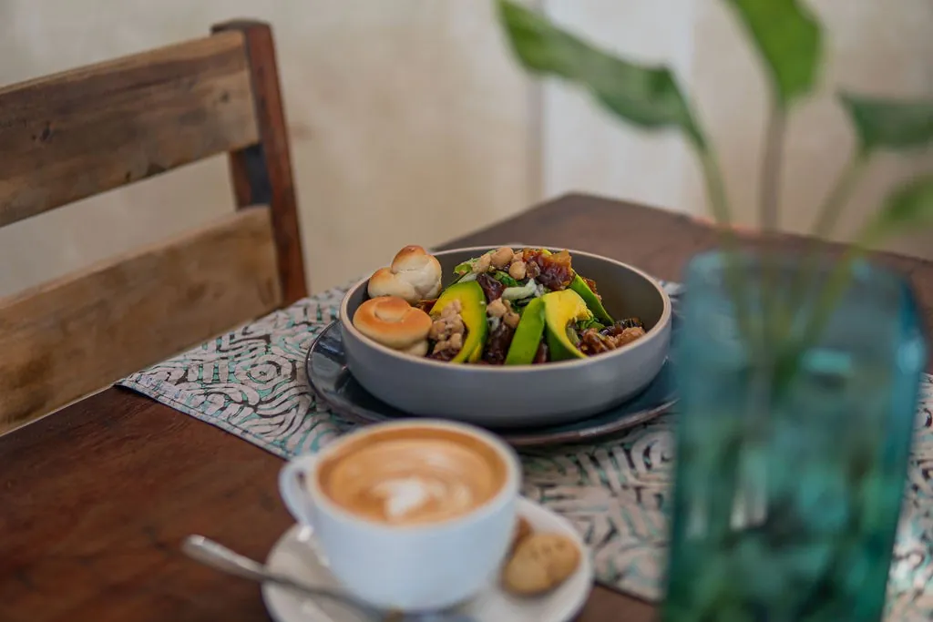 A breakfast bowl and a cappuccino with latte art on a wooden table, leafy plants behind.