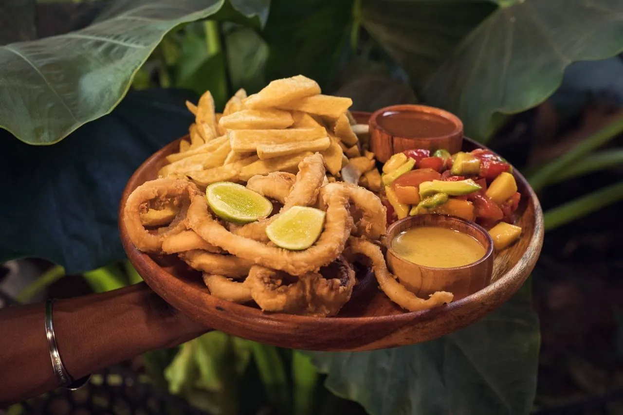 A wooden plate of fried calamari and chips with mango salsa and sauces, held out among leaves