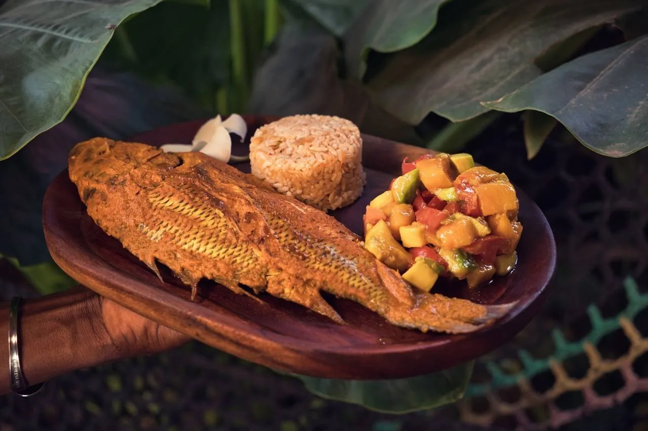 A whole spiced fried fish with a rice cake and mango-avocado salad on a wooden plate