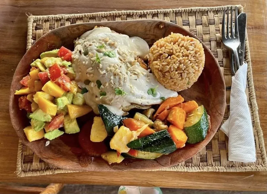 A wooden plate with grilled vegetables, mango-avocado salad, a rice cake and a creamy topping, on a woven mat