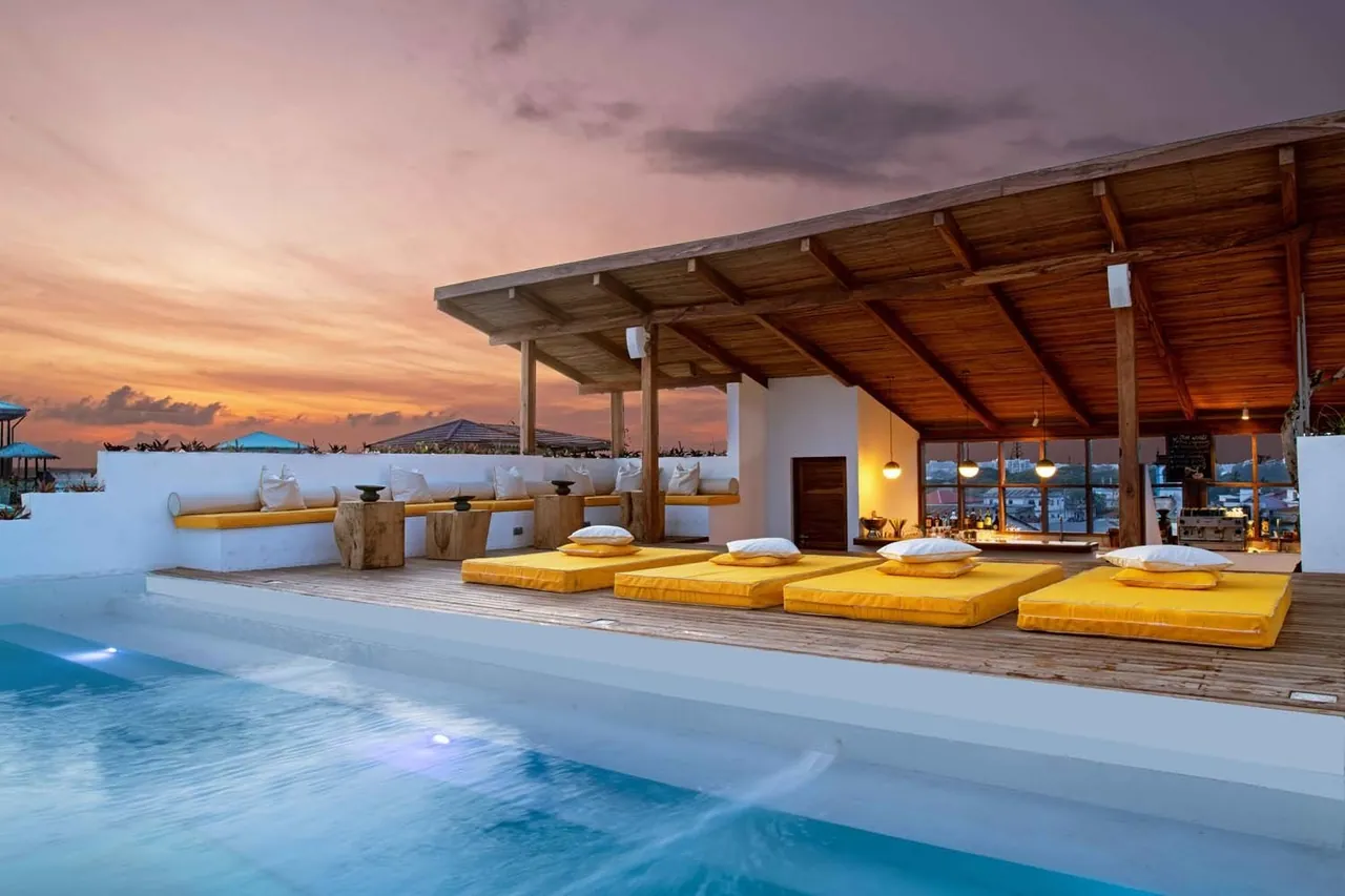 A rooftop deck with yellow loungers beside a pool under a pink sunset sky, a covered bar behind