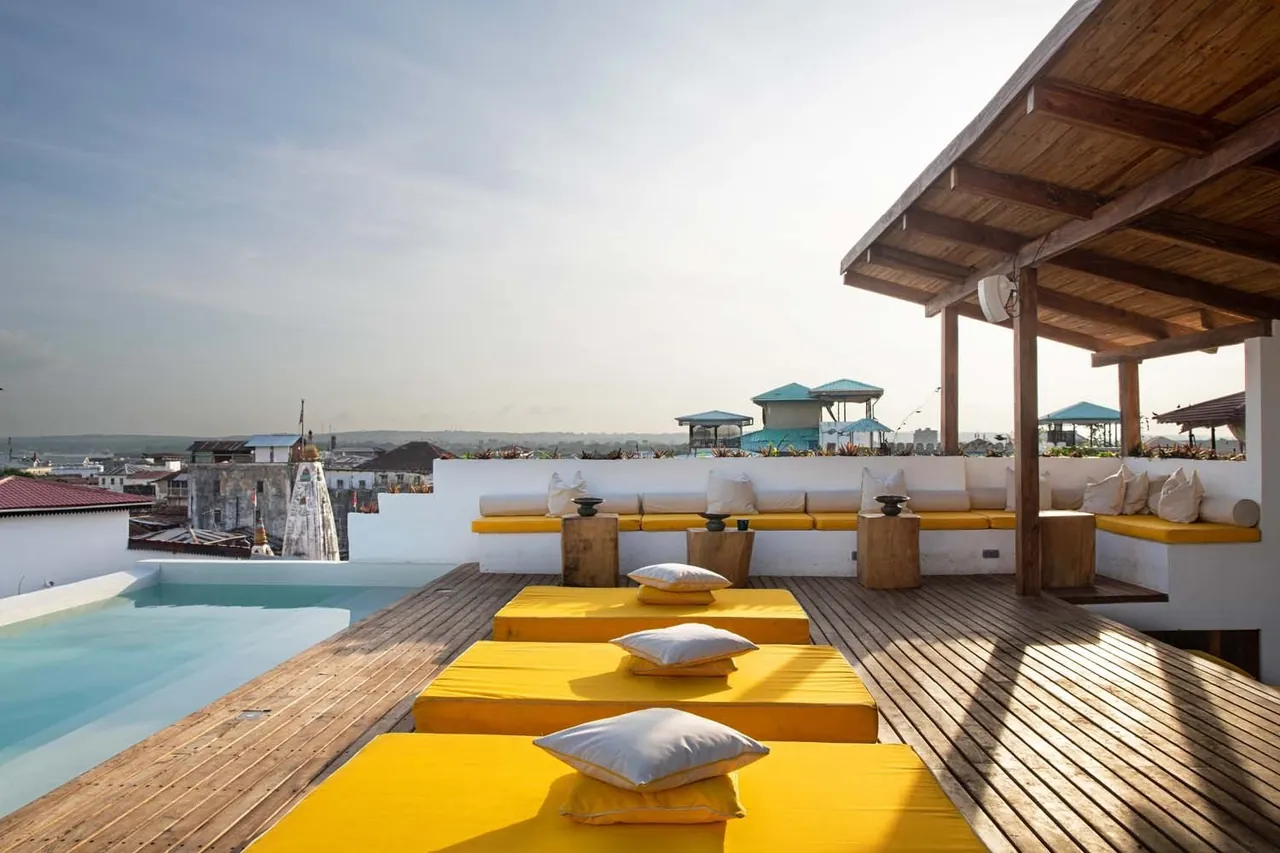 A wooden rooftop deck with yellow cushions and a lounge, overlooking Stone Town and the harbour