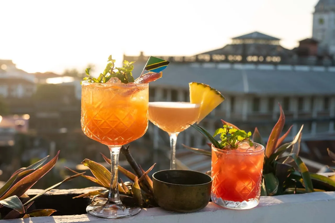 Three orange cocktails garnished with pineapple and herbs, Stone Town rooftops behind